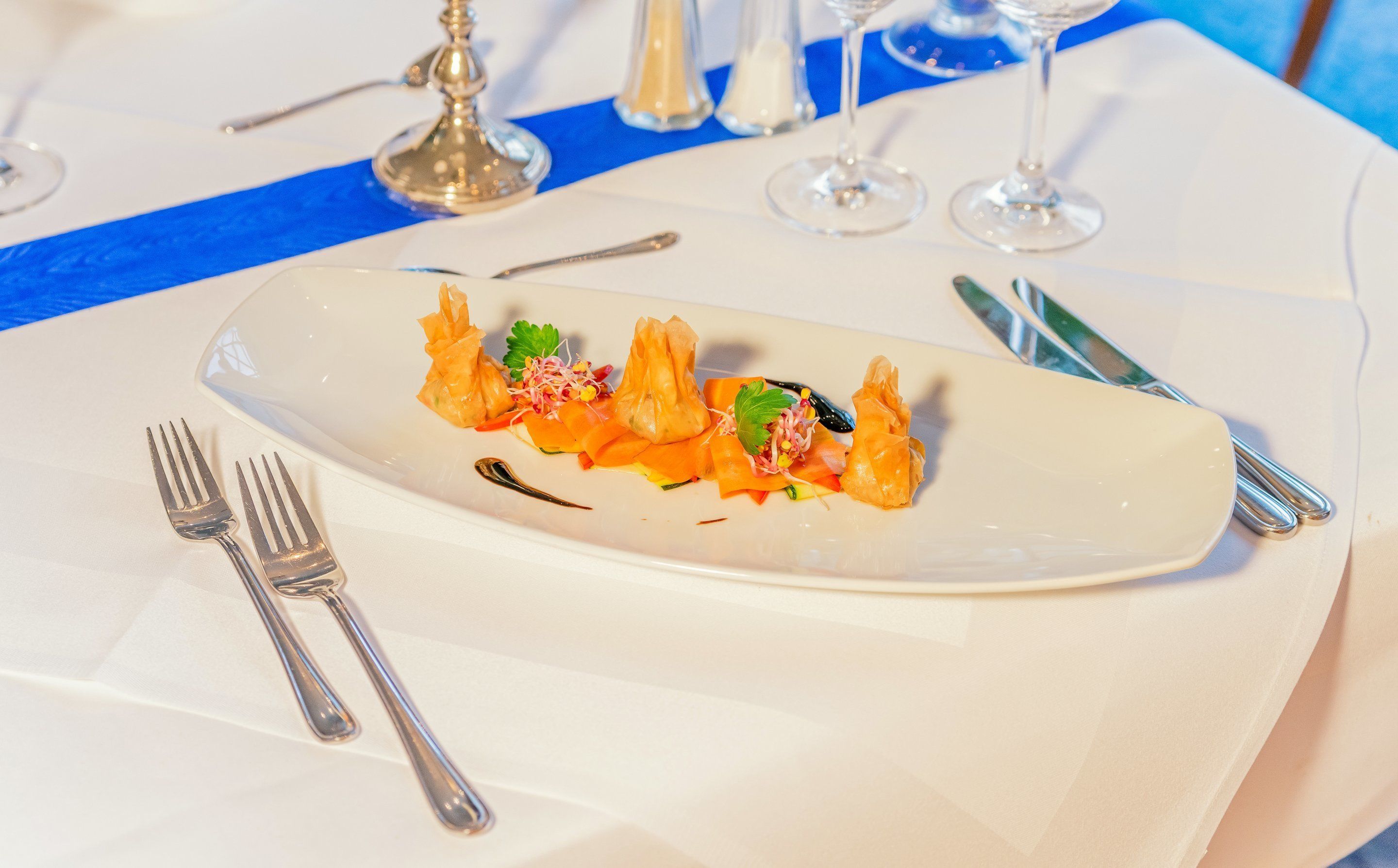 Ein weißer, ovaler Teller mit einem Fischgericht, dazu Besteck und eine blaue Serviette auf einem Tisch mit weißer Tischdecke.