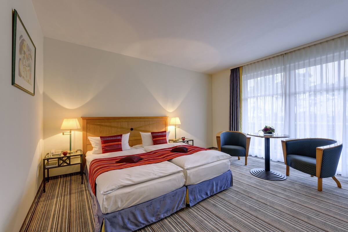 A hotel room with a bed, two bedside tables, a lamp, a chair, a small table, and a window with curtains.