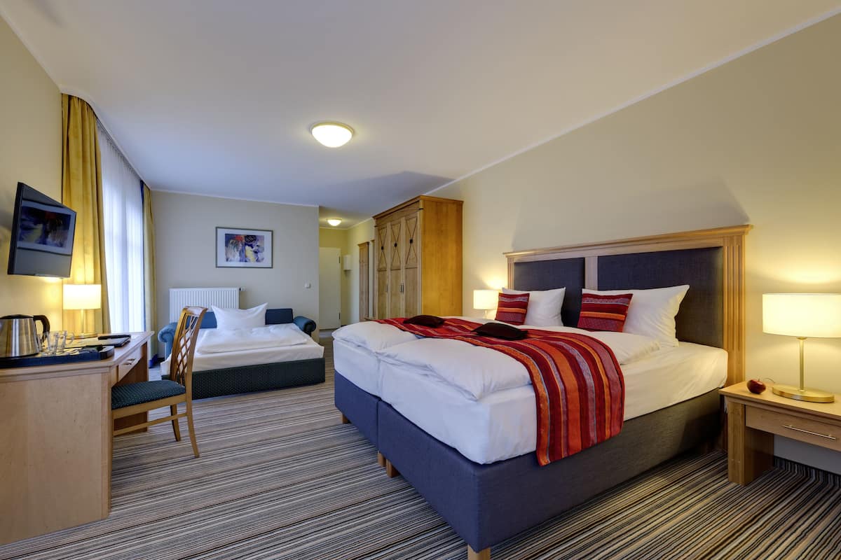A hotel room with a large bed, a smaller bed, a desk with a chair, a television, and a wardrobe.