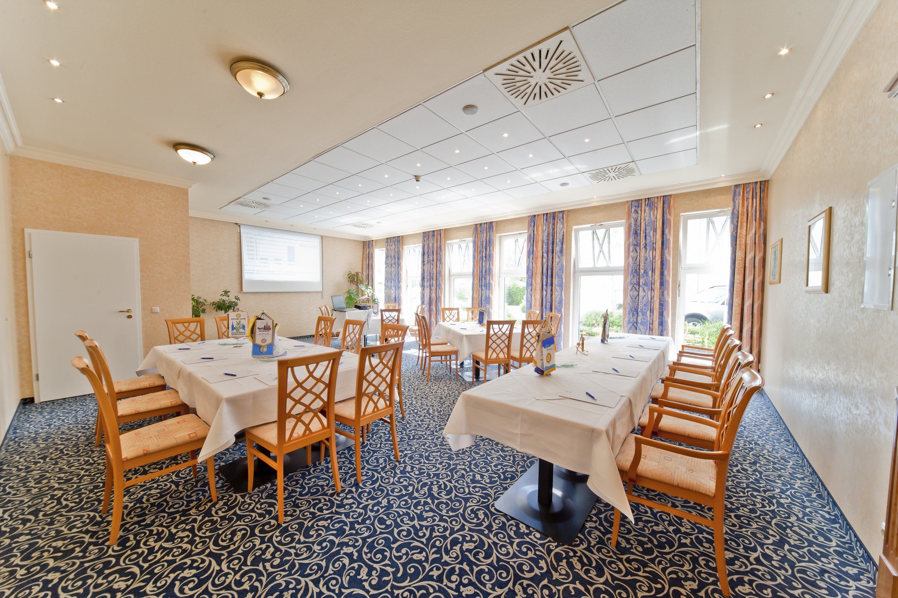 A conference room with multiple tables and chairs, a patterned carpet, and large windows with curtains.