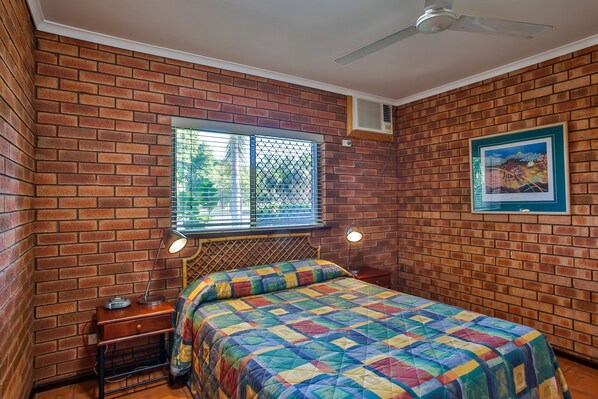 A bedroom with brick walls, a bed with a colorful quilt, a window with metal bars, a ceiling fan, and a framed picture on the wall. - Bayside Holiday Apartments (Broome)