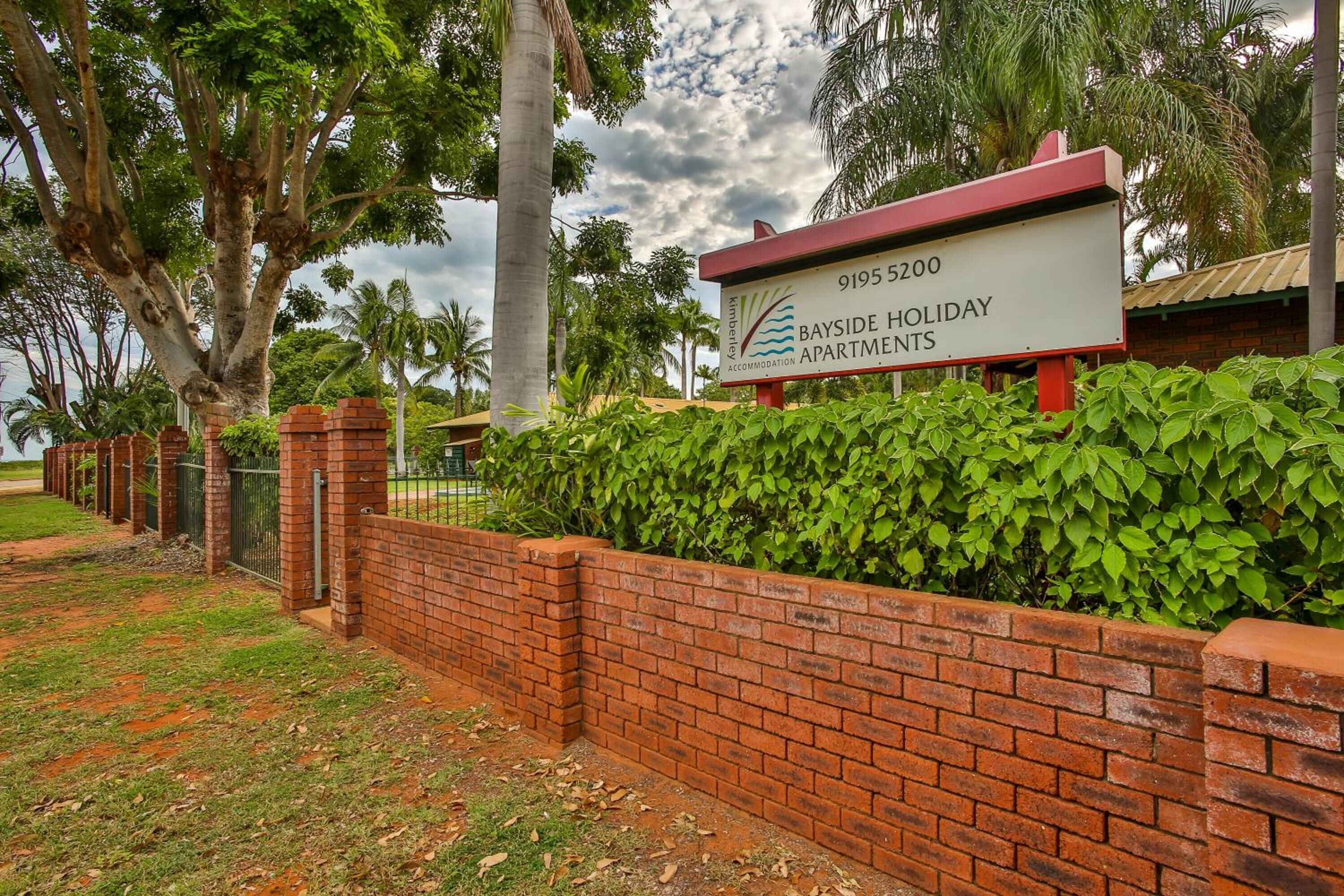 A sign for Bayside Holiday Apartments with a brick wall and greenery in the background.