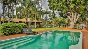 Una piscina con sillones de descanso, rodeada de abundante vegetación y un edificio pequeño.