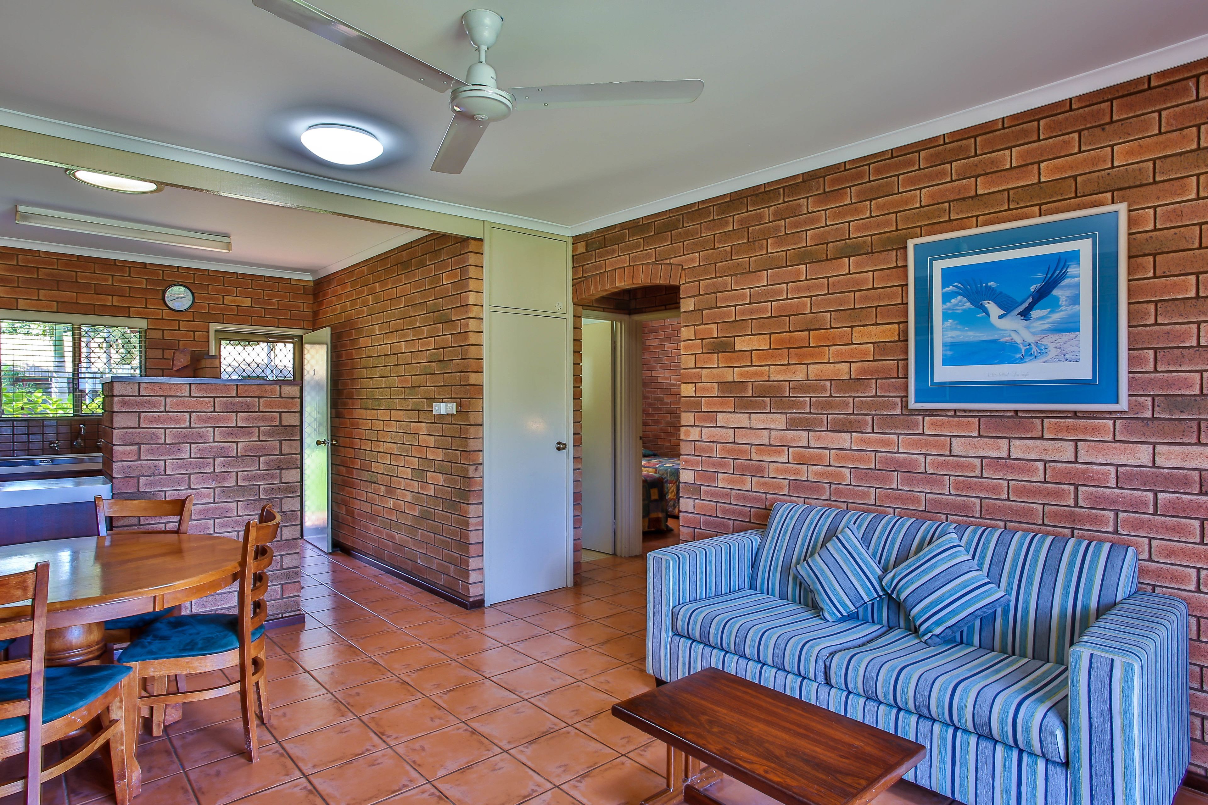 A living room with a brick wall, a striped sofa, a wooden dining table with chairs, and a ceiling fan.