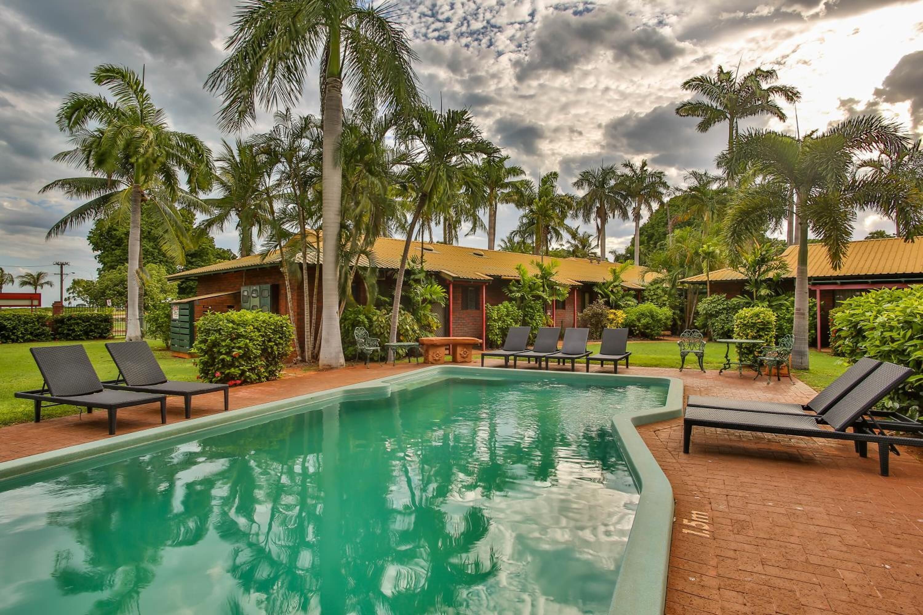 A swimming pool with lounge chairs, surrounded by palm trees and a building with a tiled roof.