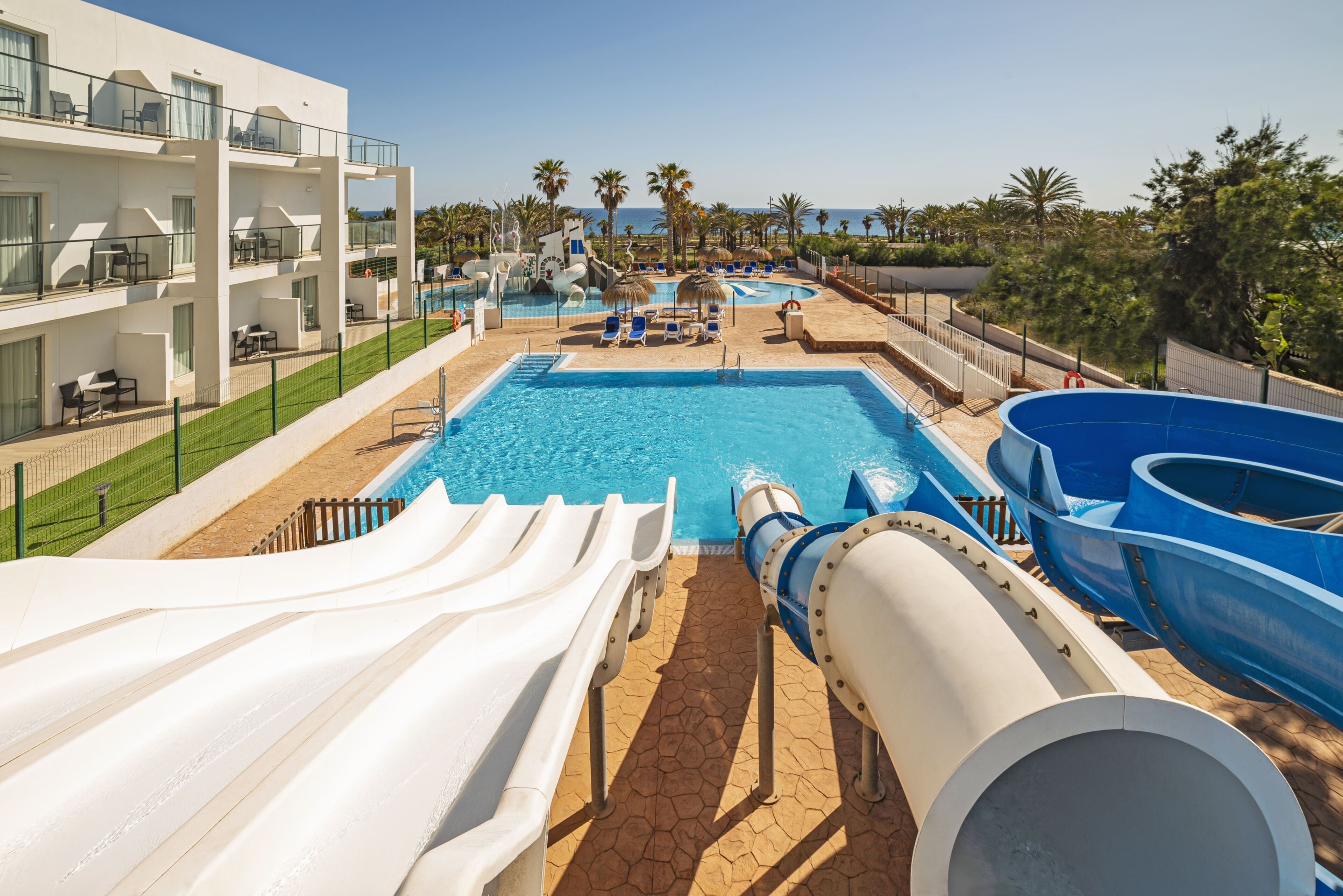 Un área de piscina con toboganes y sillas de descanso, piscina y un edificio con balcones.