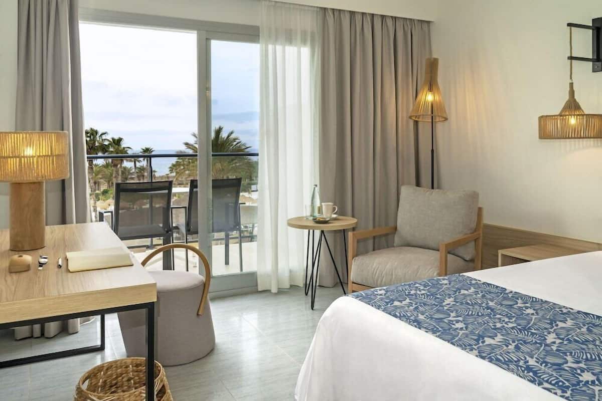Ein Hotelzimmer mit Bett, Schreibtisch, Stuhl, kleinem Tisch und einem Balkon mit Blick auf Palmen und das Meer.