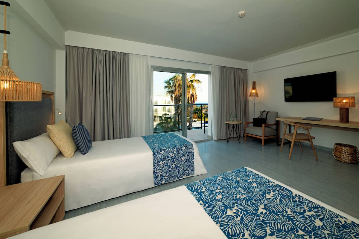 Ein Hotelzimmer mit zwei Betten, einem Schreibtisch, einem Sessel, einem Fernseher und Blick auf Palmen.