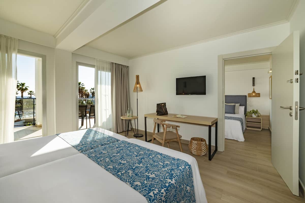 Ein Hotelzimmer mit einem großen Bett, einem Schreibtisch, einem Stuhl, einem Fernseher und einem Balkon mit Blick auf Palmen.