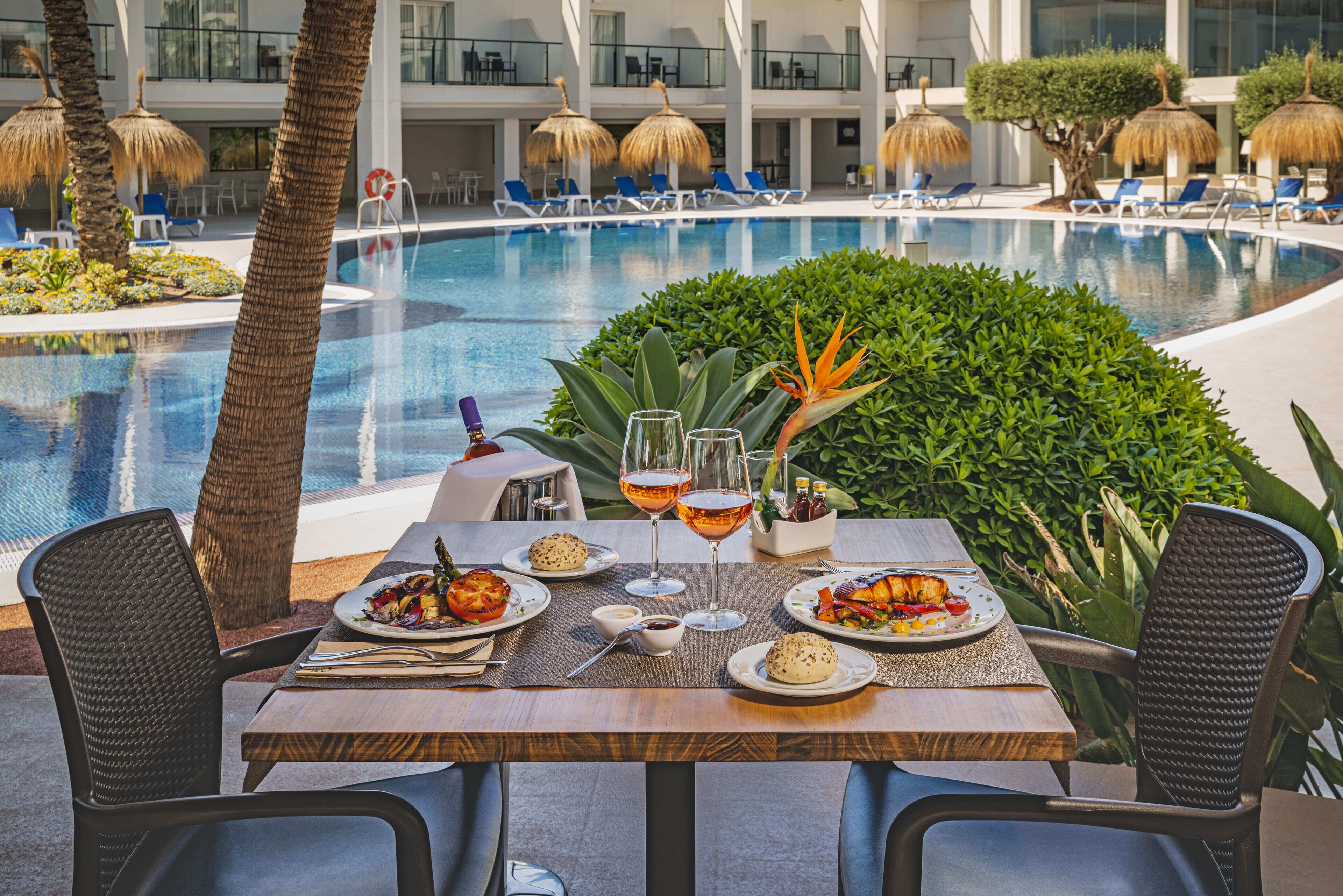Una mesa de comedor con platos de comida y copas de vino, con vistas a una piscina con sillones y sombrillas.