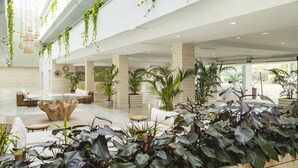Un vestíbulo amplio con una zona de estar central, plantas en macetas y un techo alto con luces colgantes.
