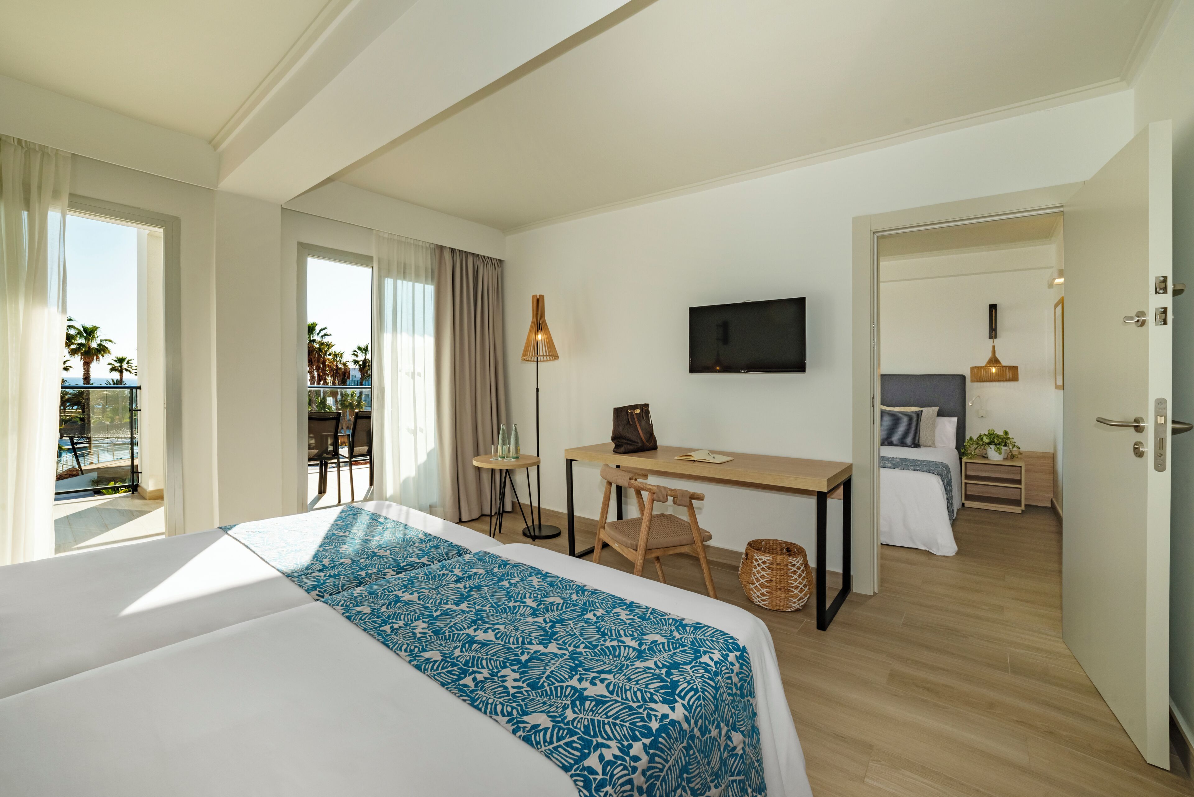 A hotel room with a large bed, a desk, a chair, a TV, and a balcony with a pool view.