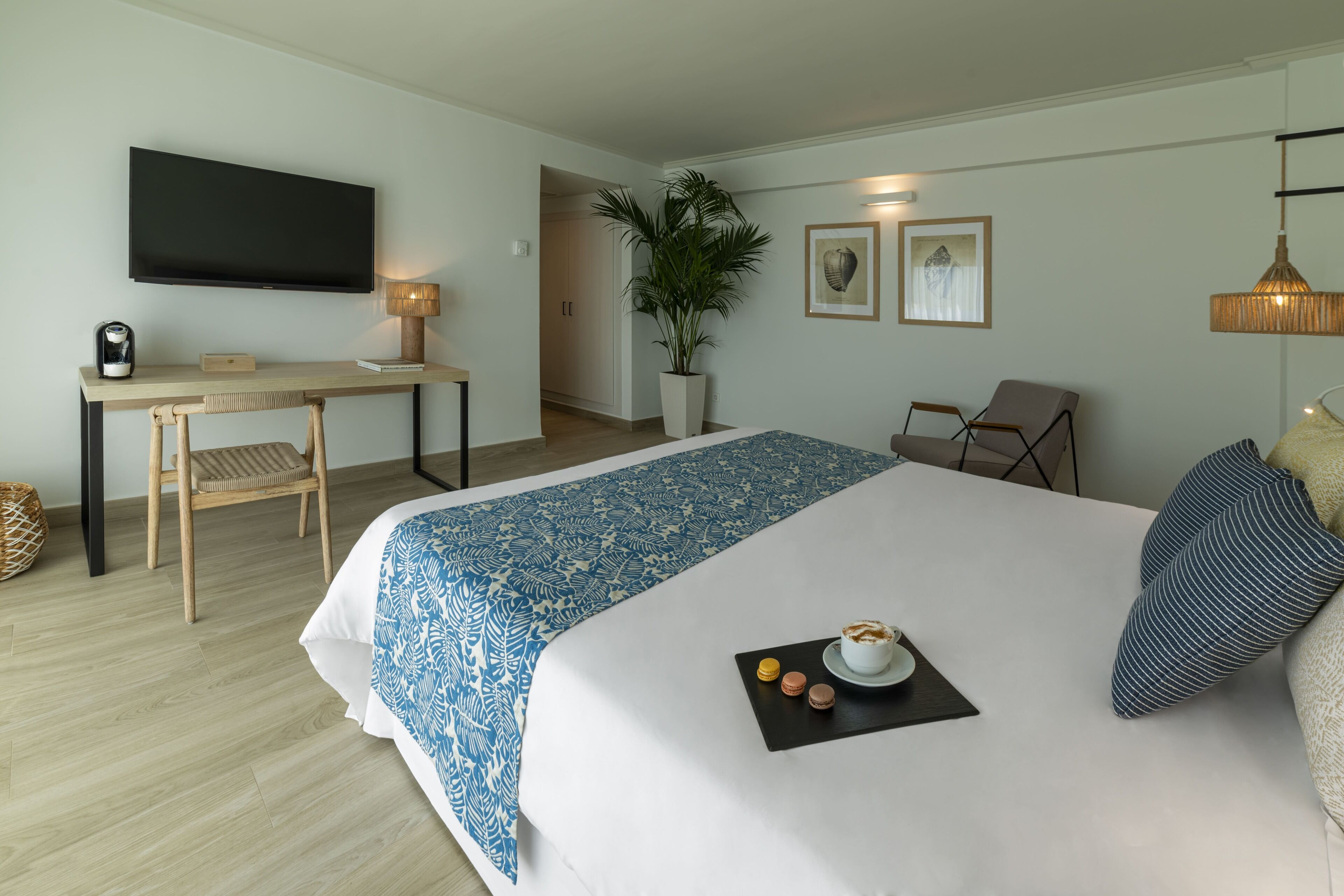 A hotel room with a large bed, a desk with a chair, a television, a potted plant, and a dining area with a plate of food.