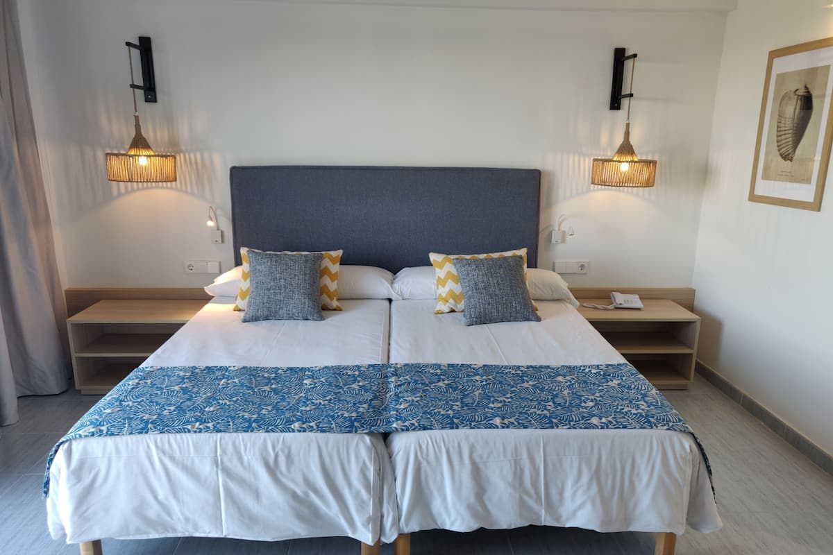 Ein Doppelbett mit einer blau-weißen Bettdecke, zwei Kissen mit Zickzackmuster und ein grauer Kopfteil. Dazu gehören Nachttische mit Lampen, ein gerahmtes Bild an der Wand und ein Nachttisch aus Holz.
