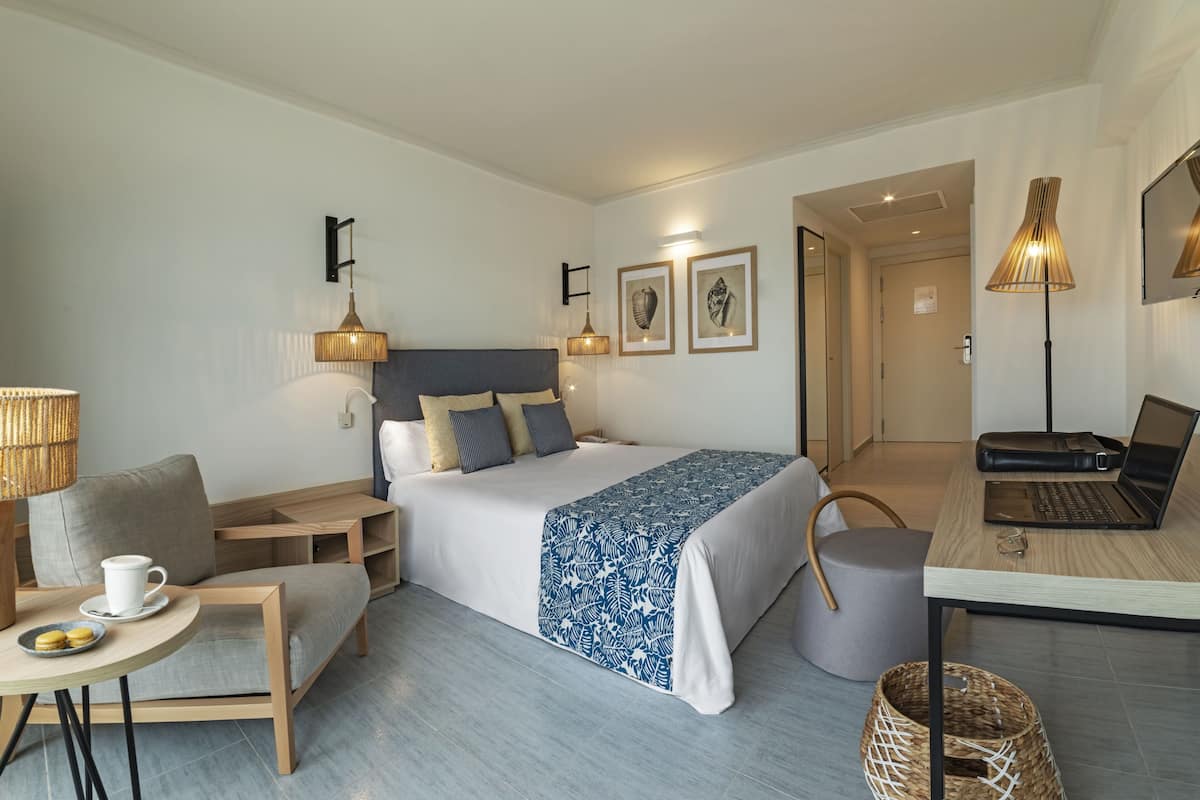 Ein modernes Hotelzimmer mit einem Bett, Nachttischen, einem Schreibtisch mit einem Laptop, einem Sessel und einem kleinen Tisch mit Tasse und Keksen.