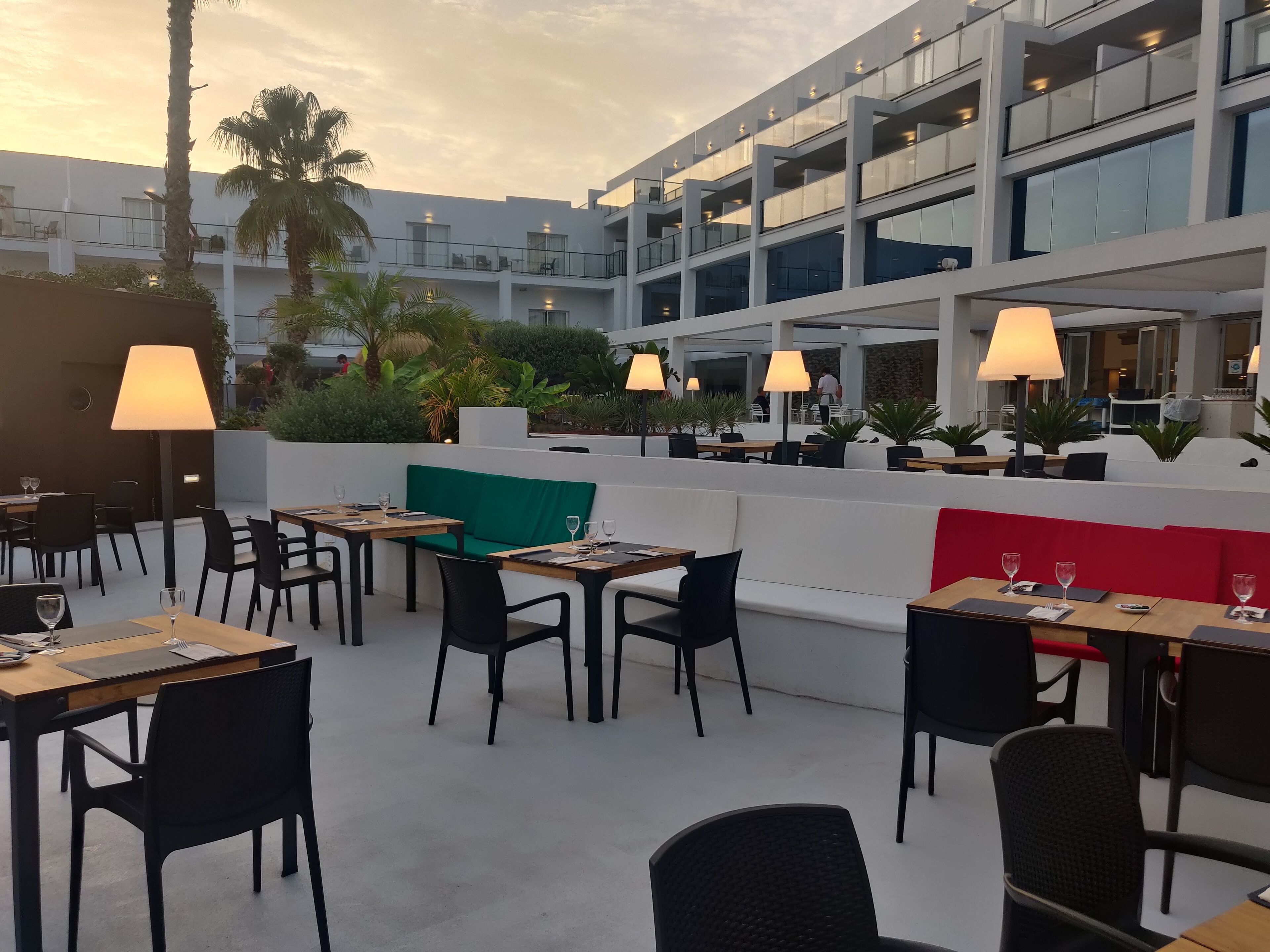 An outdoor dining area with tables and chairs, surrounded by greenery and a modern building with balconies.