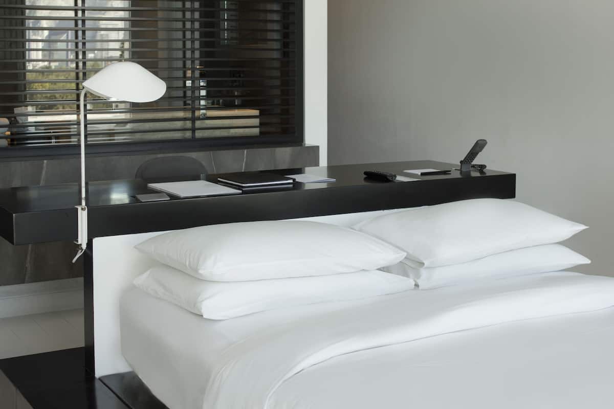A modern bedroom with a bed, white pillows, a bedside table with a lamp, and a window with blinds.