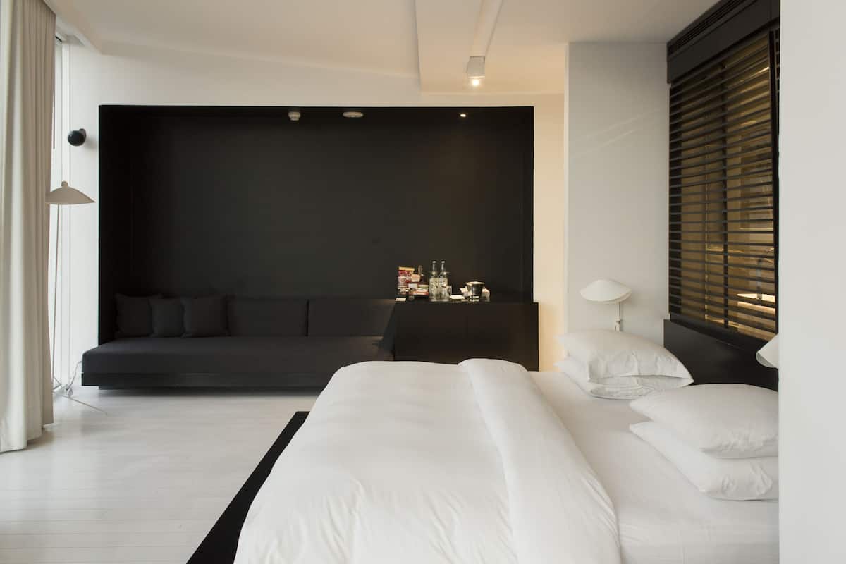 A modern bedroom with a large bed, a dark accent wall, a sofa bed, a bedside table with a lamp, and a window with blinds.