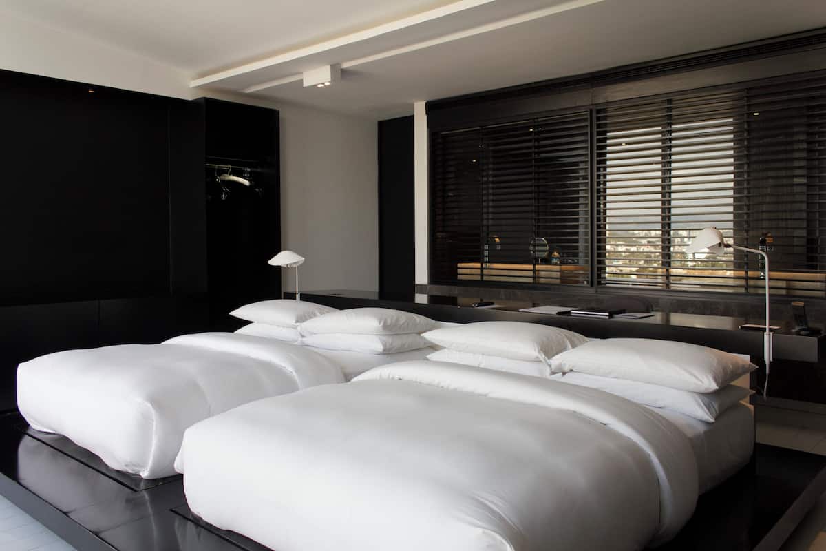 A modern bedroom with a large bed, a bedside table, a lamp, and a window with blinds.