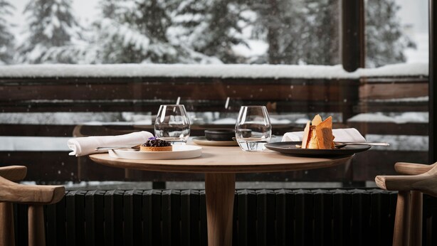 A dining table with plates, glasses, and a napkin, set against a snowy landscape with trees. - Grand Hôtel & Spa NUXE Serre Chevalier (Saint-Chaffrey)