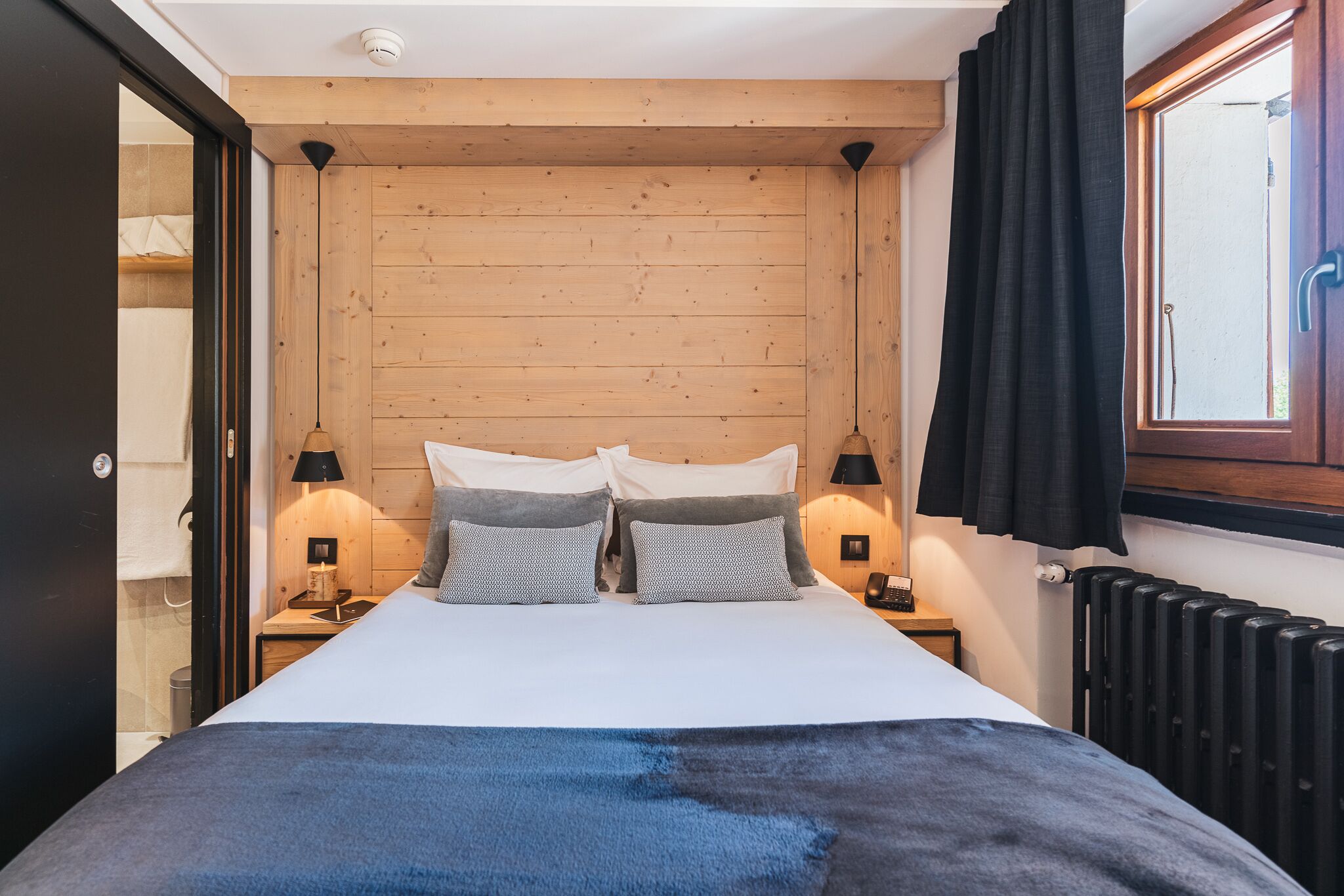 Ein Schlafzimmer mit einem hölzernen Kopfteil, zwei Nachttischen mit Lampen, einem Bett mit blauer Decke und einem Fenster mit schwarzen Vorhängen.