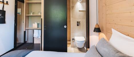 A modern hotel room with a bed, a wooden wardrobe, a bathroom with a toilet and sink, and a small shelf with toiletries.