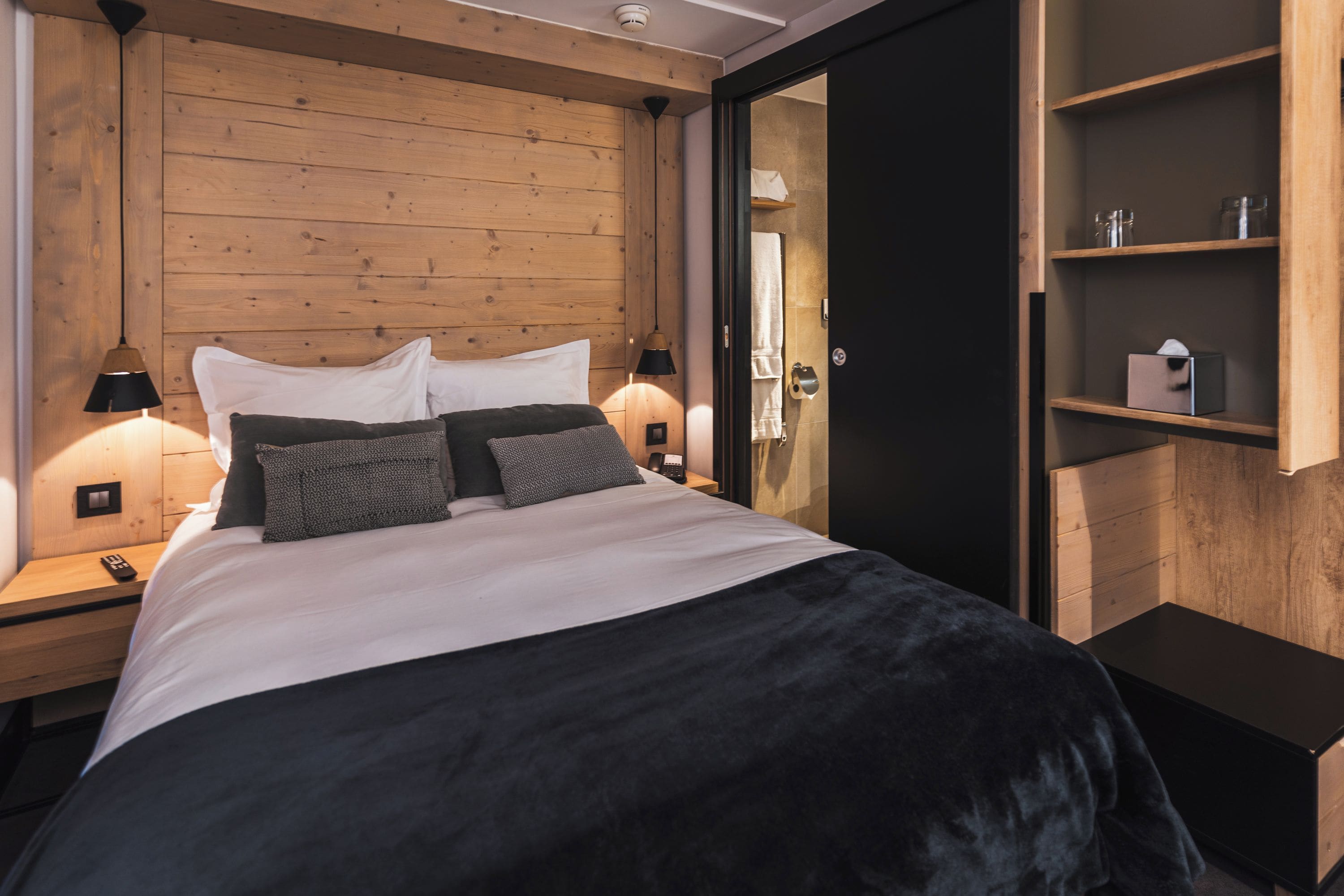 A modern bedroom with a wooden headboard, a large bed with multiple pillows, bedside lamps, and a built-in shelf with toiletries.