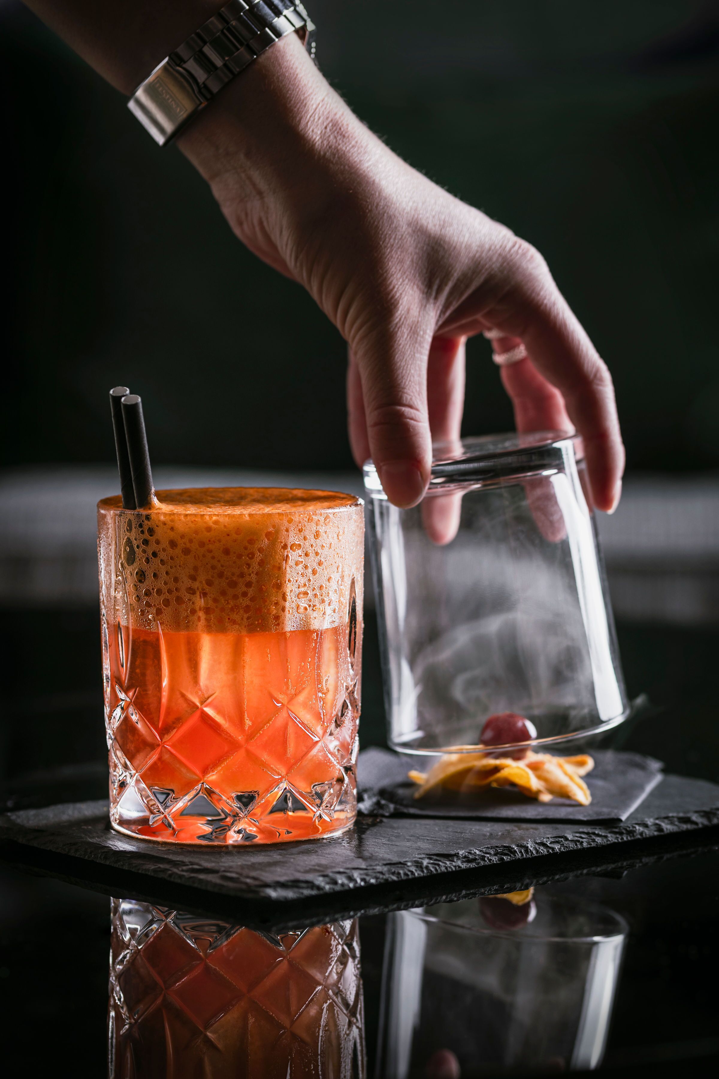 Une main tenant un verre avec une paille, à côté d’un verre transparent contenant une tranche de citron et un petit zeste d’orange, le tout posé sur une surface noire.