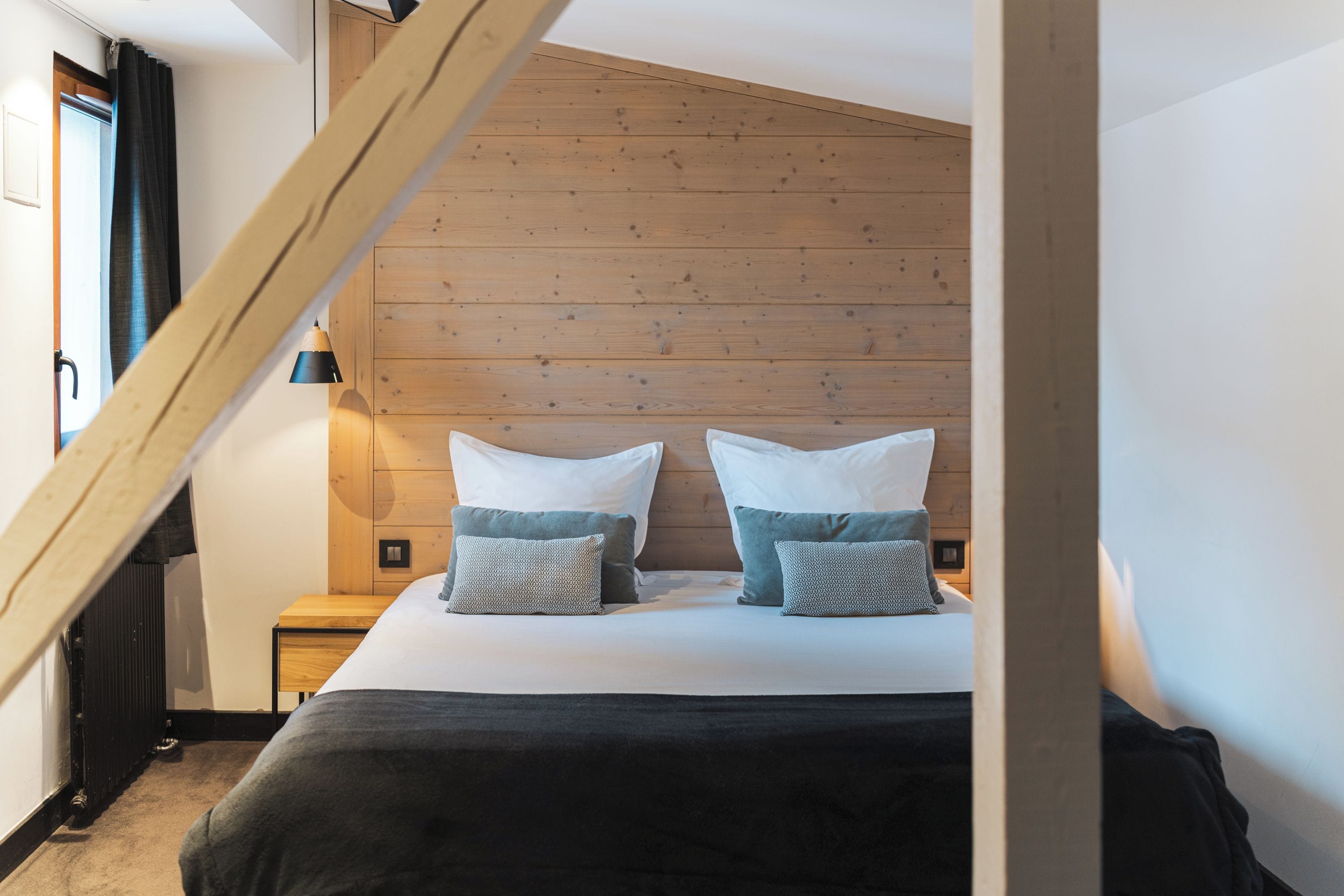 Un dormitorio con techo inclinado, una cama con ropa de cama blanca y almohadas estampadas, una mesita de noche con lámpara y cabecero de madera.