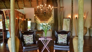 A lobby with two leather armchairs, a wooden table, a chandelier, and a stone wall. - Imbali Safari Lodge (Imbali Concession)