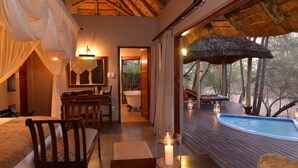 A room with a wooden desk, a chair, a bed, a bathtub, and a view of a pool and trees. - Imbali Safari Lodge (Imbali Concession)