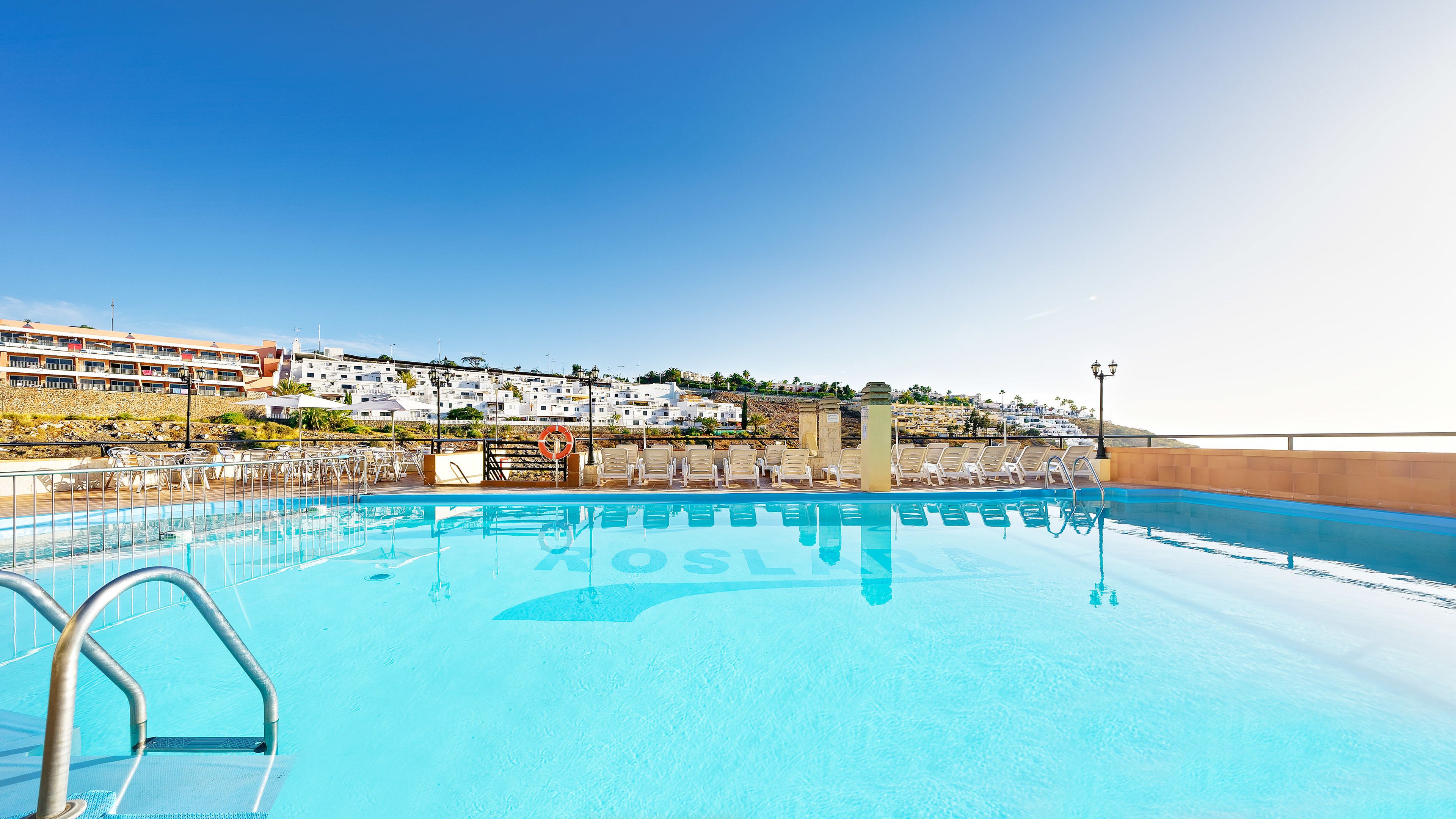 A clear blue pool with a lifebuoy, lounge chairs, and a view of a hillside with buildings.