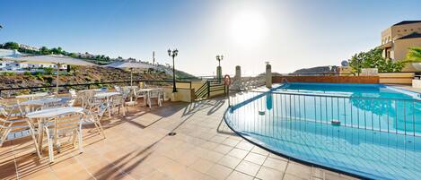 A rooftop pool area with sun loungers and tables, surrounded by a railing and overlooking a hilly landscape.