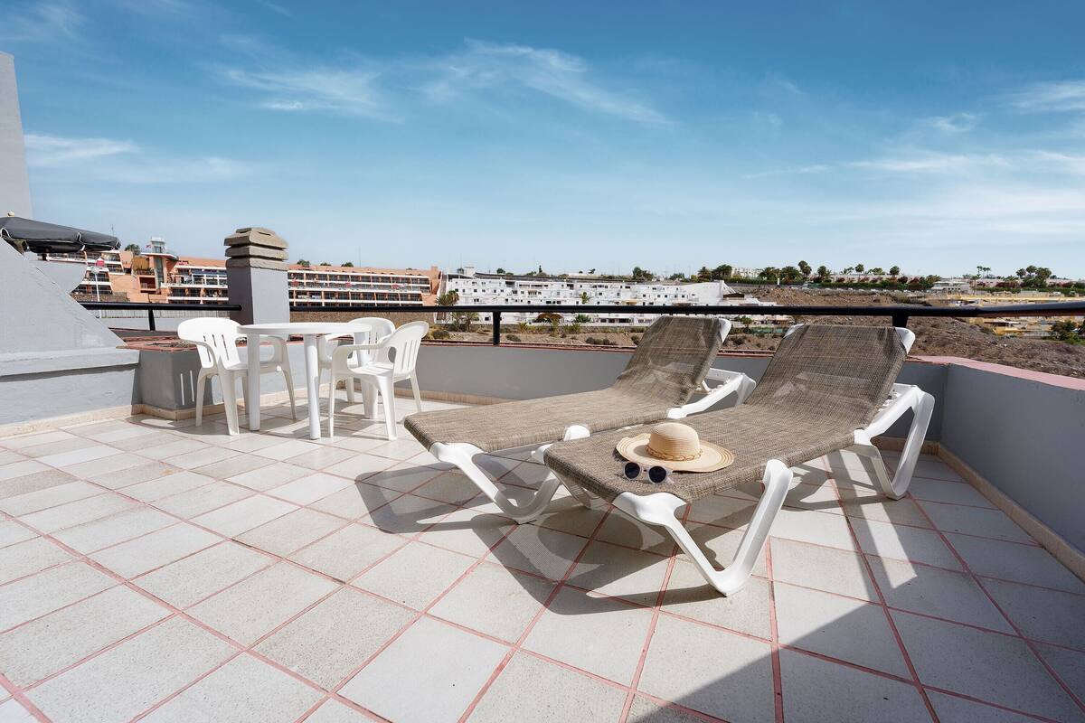 A rooftop terrace with a sun lounger, white chairs, and a table.