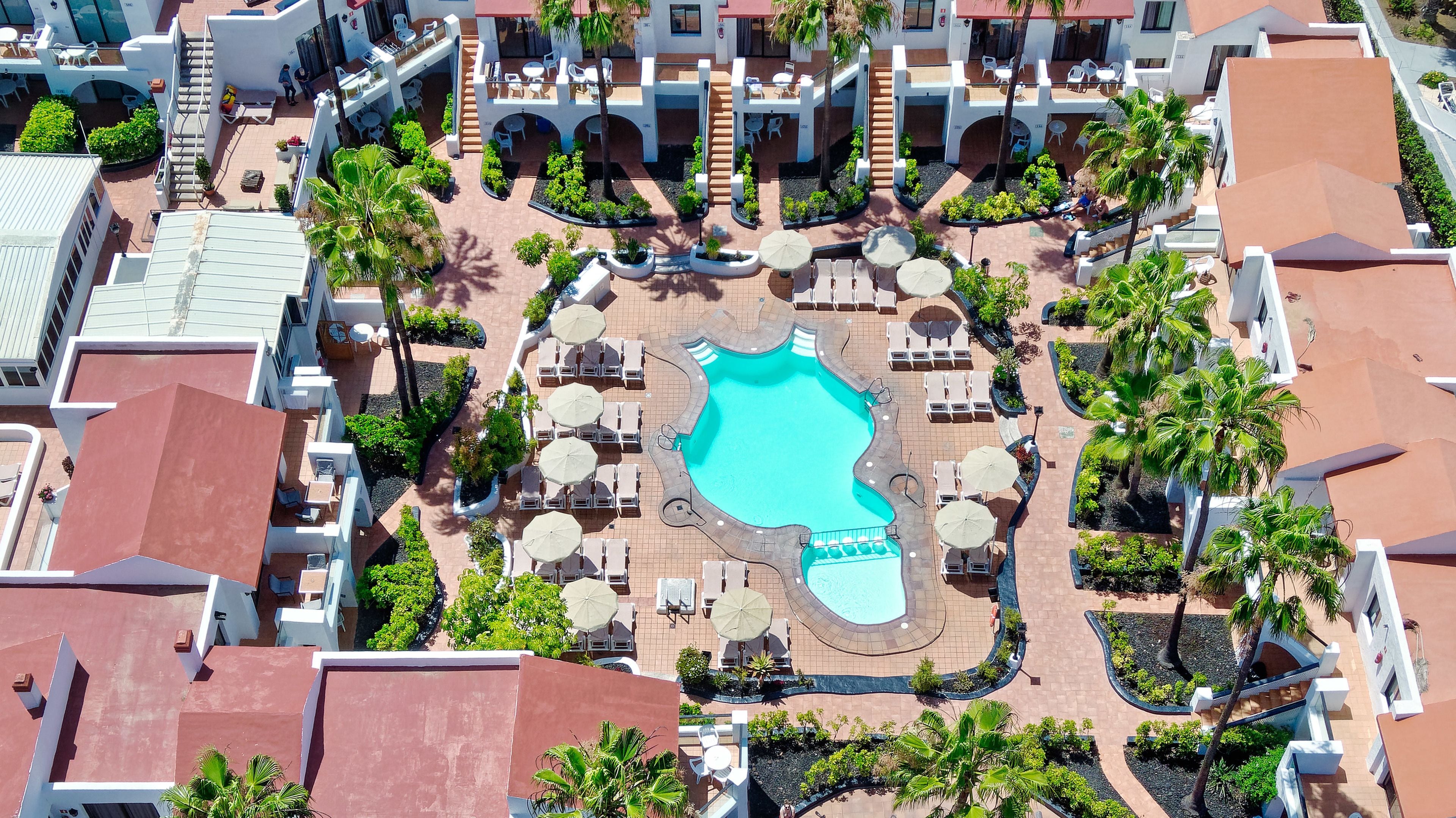 An aerial view of a resort with a central swimming pool, multiple lounge areas, and surrounding greenery.