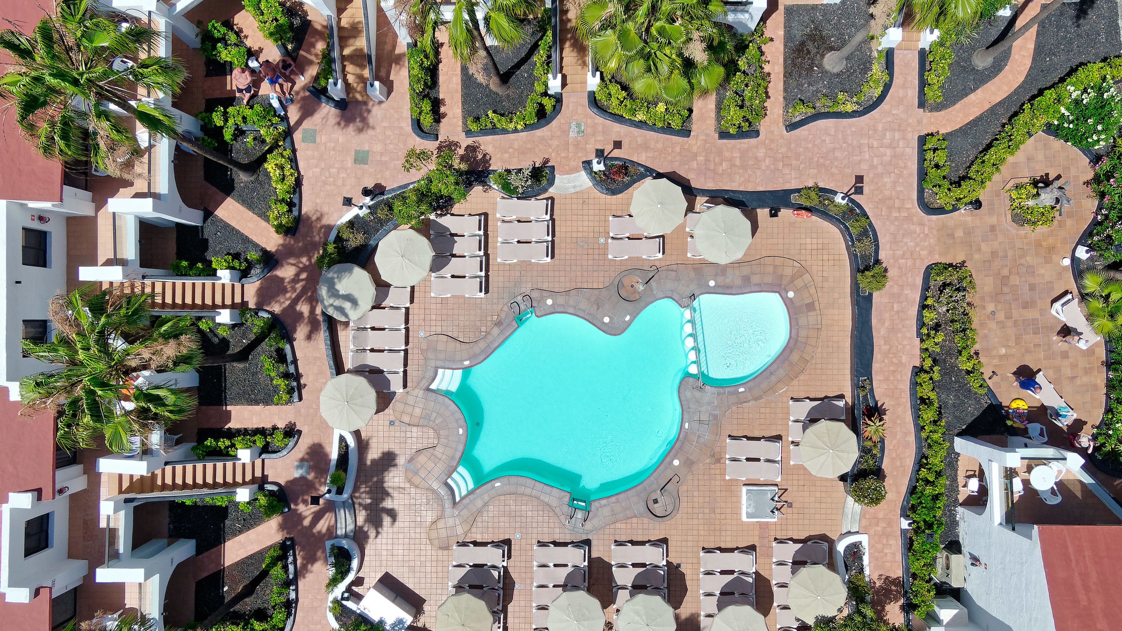 Vue aérienne d’un complexe hôtelier doté d’une piscine centrale, de chaises longues et d’une végétation luxuriante.