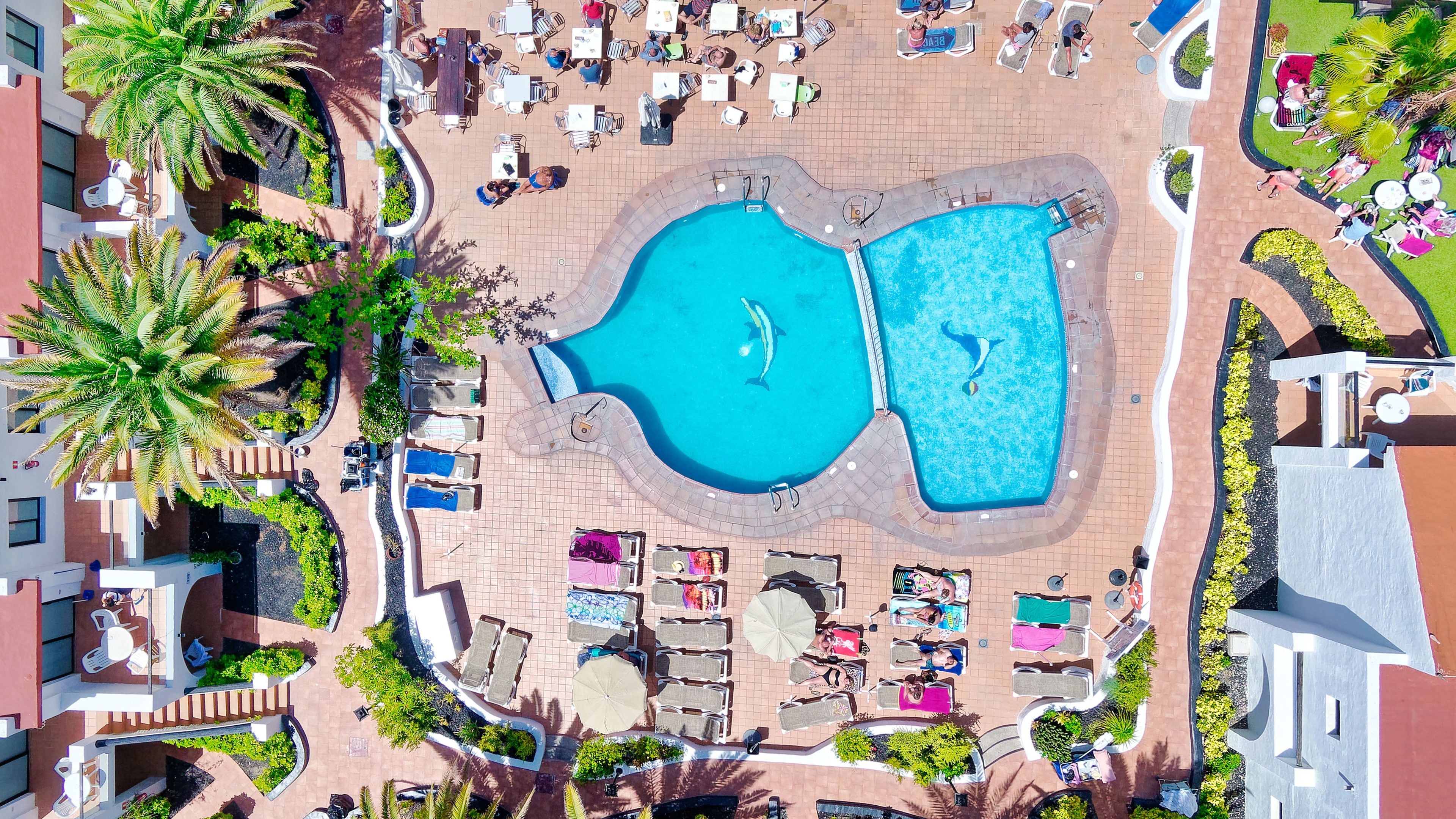 An aerial view of a resort with two swimming pools, lounge areas, and palm trees.