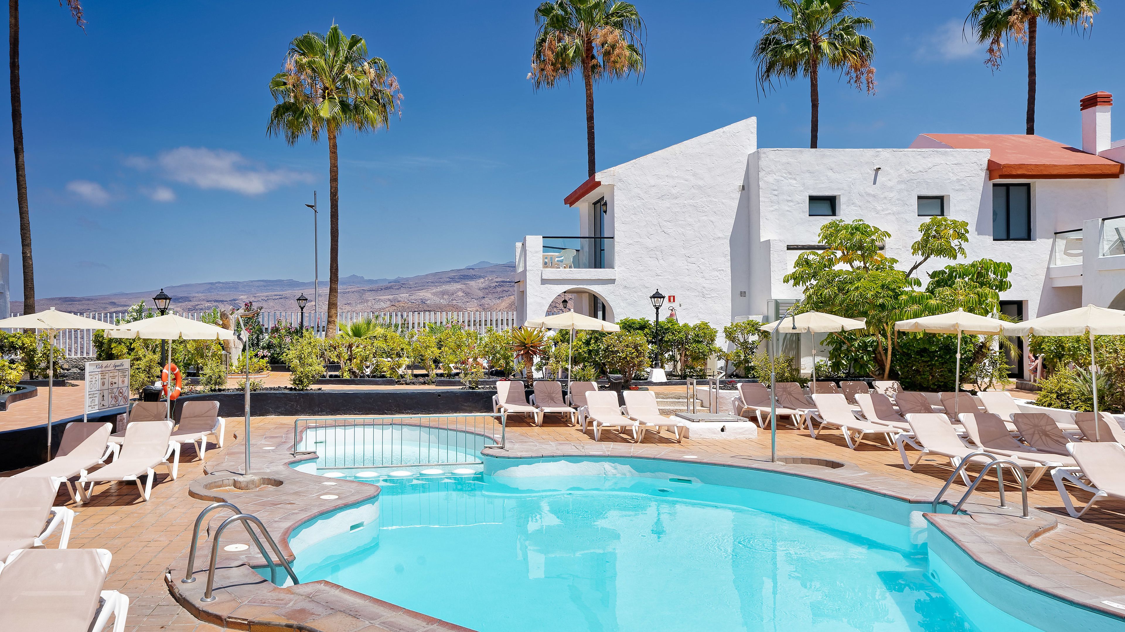 Un espace piscine agrémenté de chaises longues et de parasols, entouré de palmiers et offrant une vue sur les montagnes.