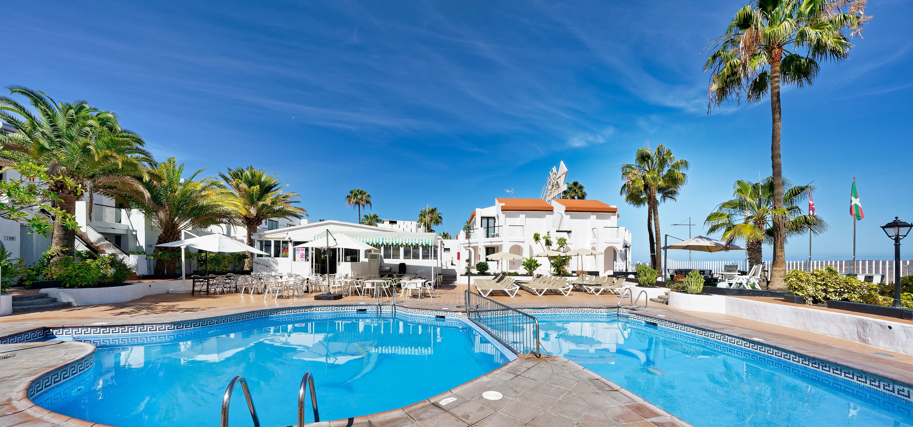 Une piscine dont le fond est recouvert de carreaux formant un motif géométrique, entourée de chaises longues et de parasols, avec un bâtiment doté d’un toit rouge en arrière-plan.