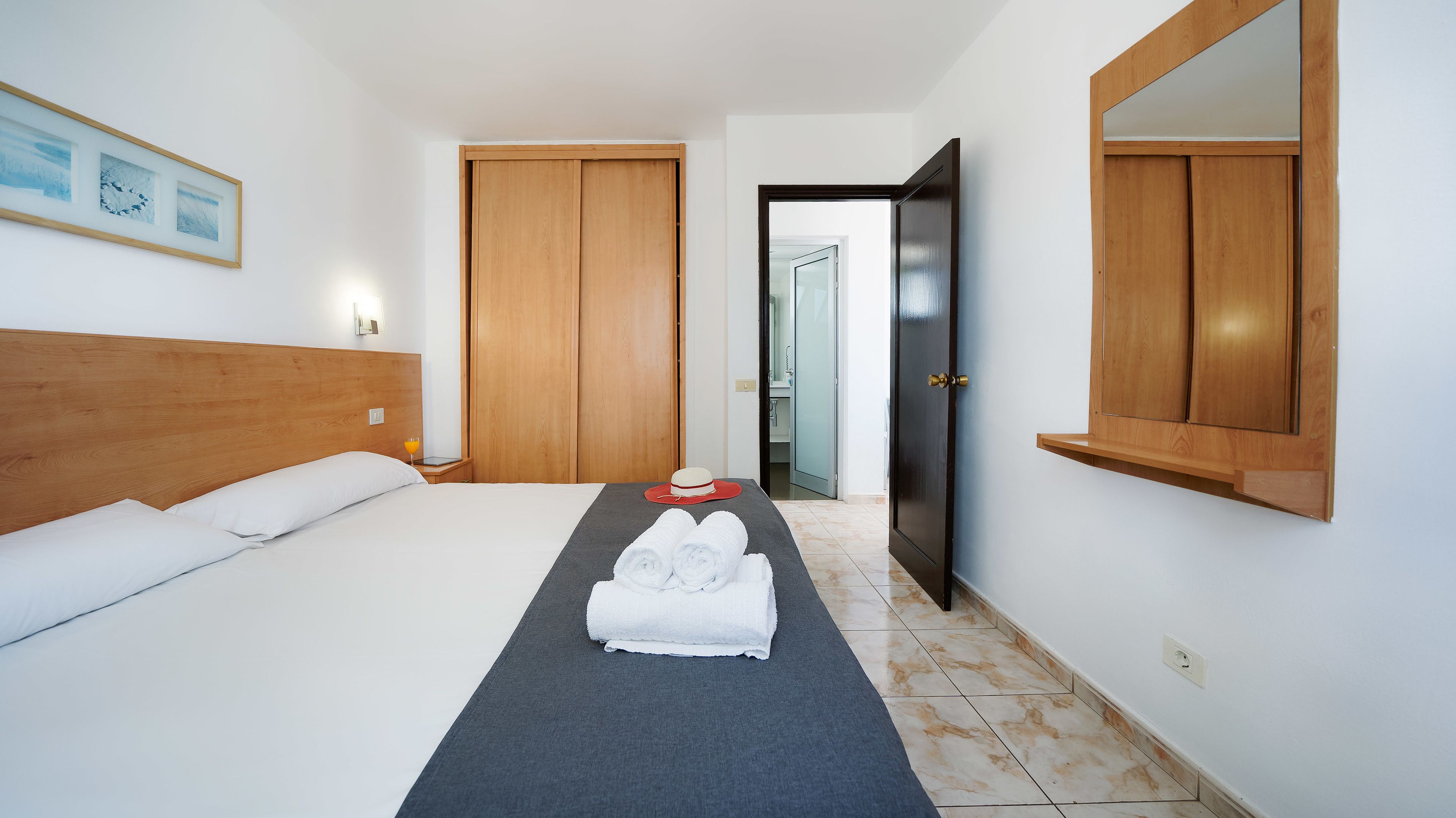 Habitación de hotel con una cama, cabecera, mesita de noche con un vaso de jugo de naranja, una toalla y un sombrero sobre la cama. Se ve una puerta de madera y un espejo en la pared.