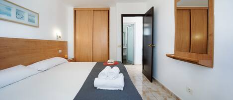 Habitación de hotel con una cama, cabecera, mesita de noche con un vaso de jugo de naranja, una toalla y un sombrero sobre la cama. Se ve una puerta de madera y un espejo en la pared.