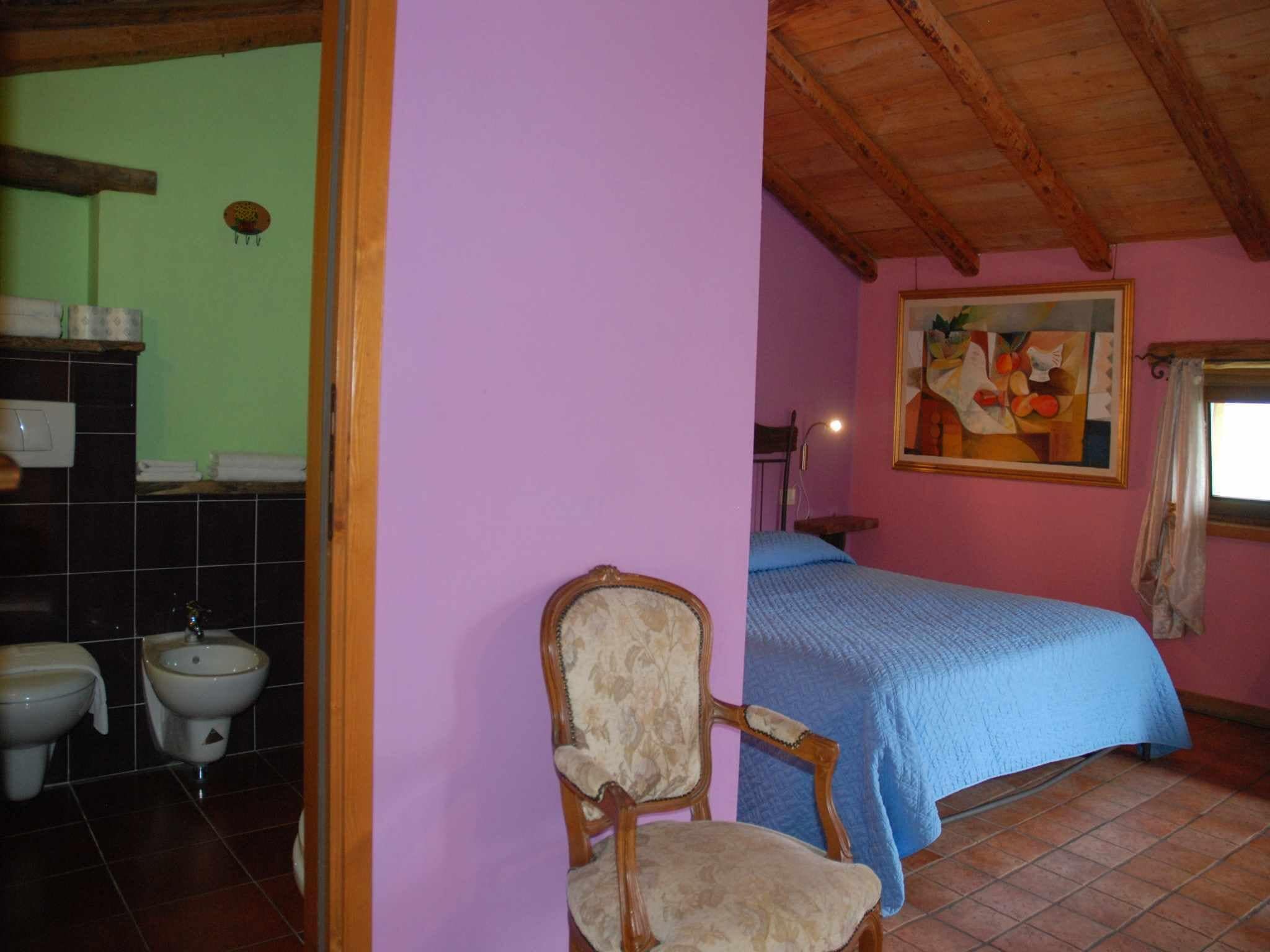 Ein Schlafzimmer mit Bett, Sessel, einem Bild an der Wand und einem Badezimmer mit Toilette und Waschbecken.