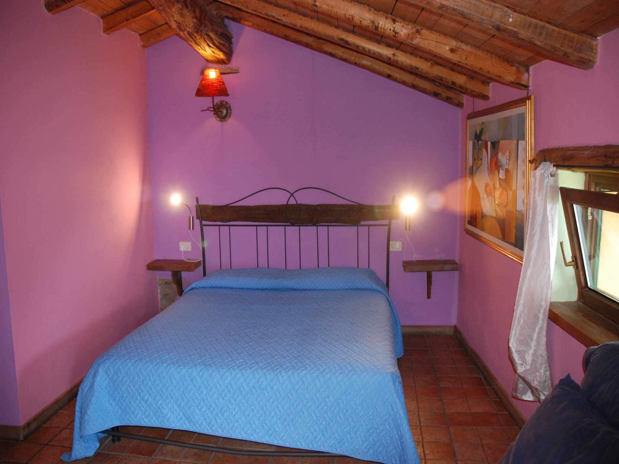 Un dormitorio con techo de madera, cama con colcha azul, dos lámparas de noche, una ventana con cortina y un cuadro en la pared.