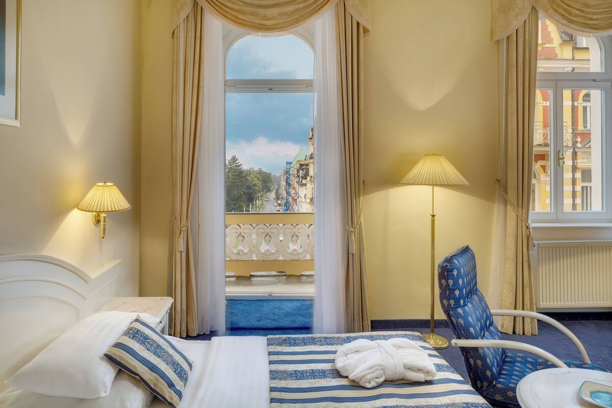 Habitación de hotel con cama, dos lámparas, una silla y vistas a un balcón con edificios.