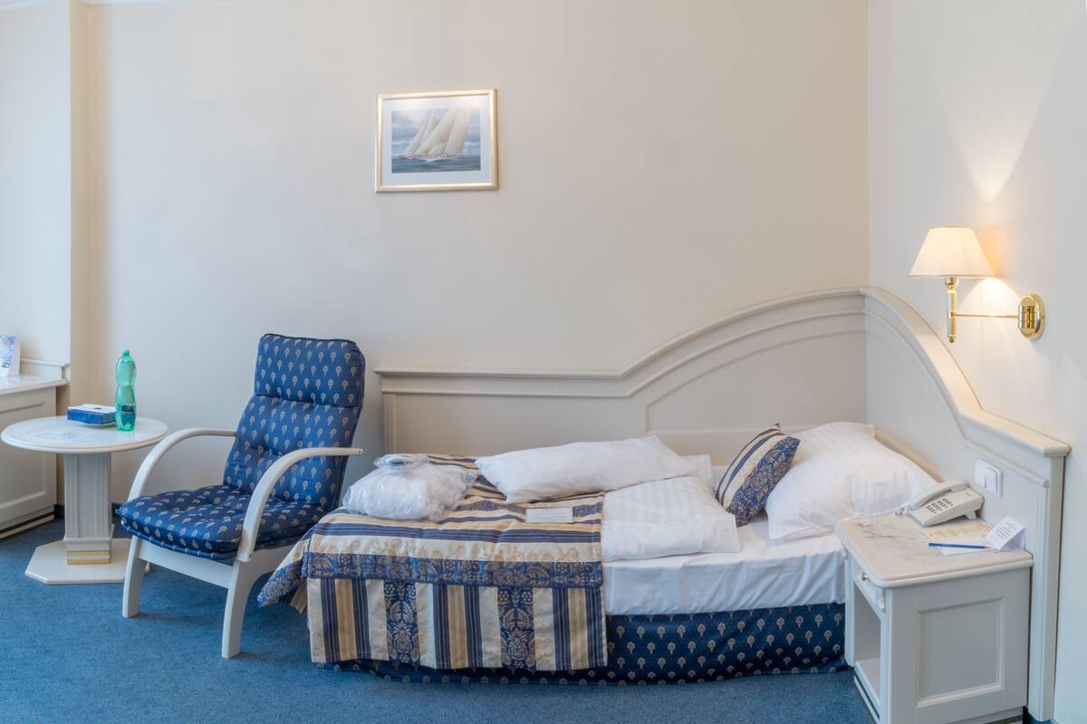 Una habitación de hotel con cama, una silla, una mesita pequeña, una lámpara de pared y un cuadro enmarcado.