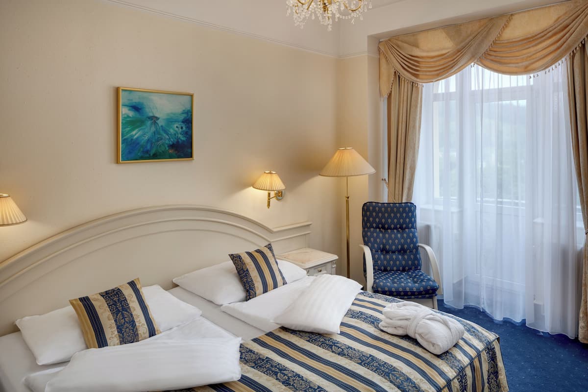 Una habitación de hotel con cama, una silla, una lámpara, un cuadro y una ventana con cortinas.
