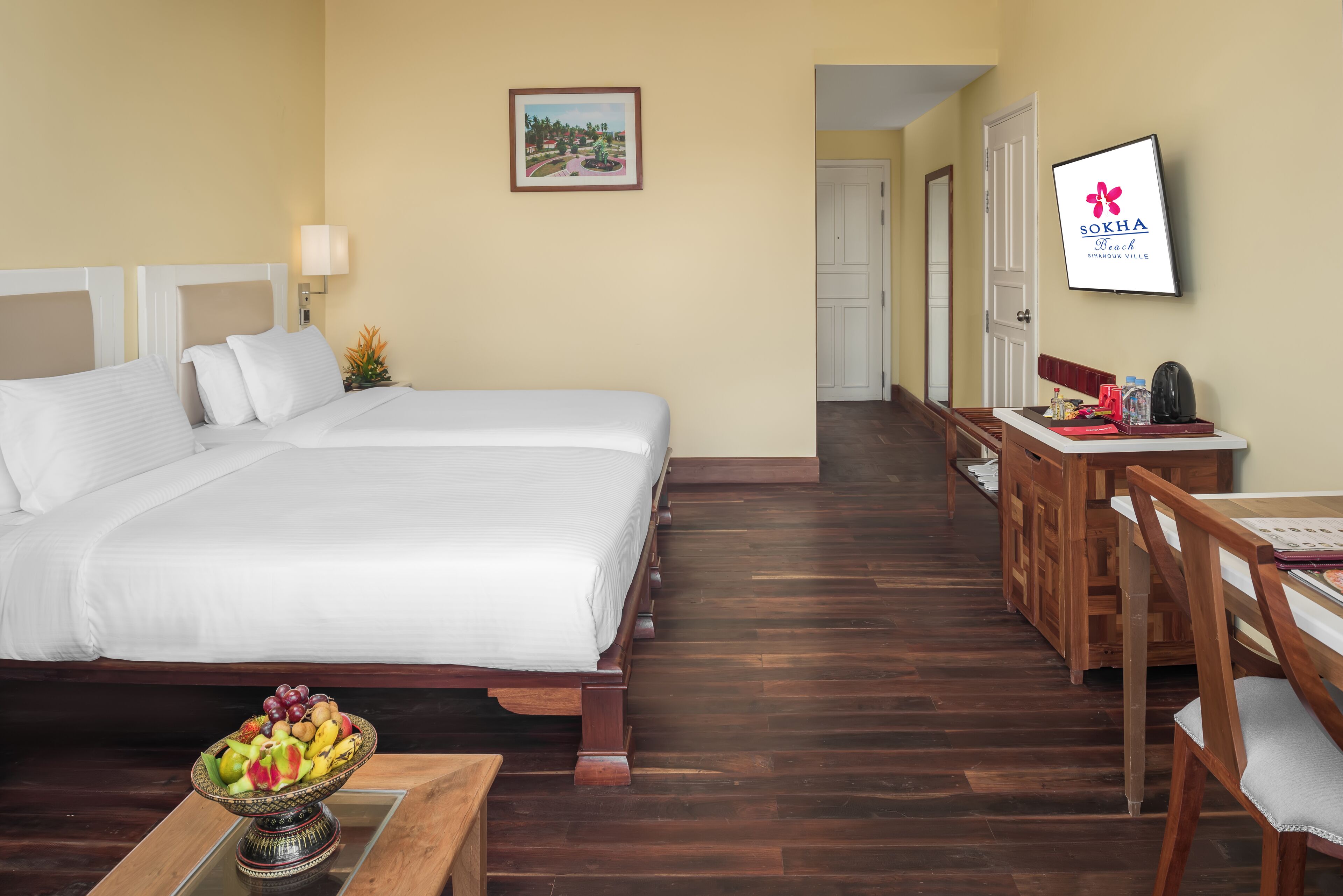 A hotel room with two beds, a dining area, a TV, and a fruit bowl on the nightstand.