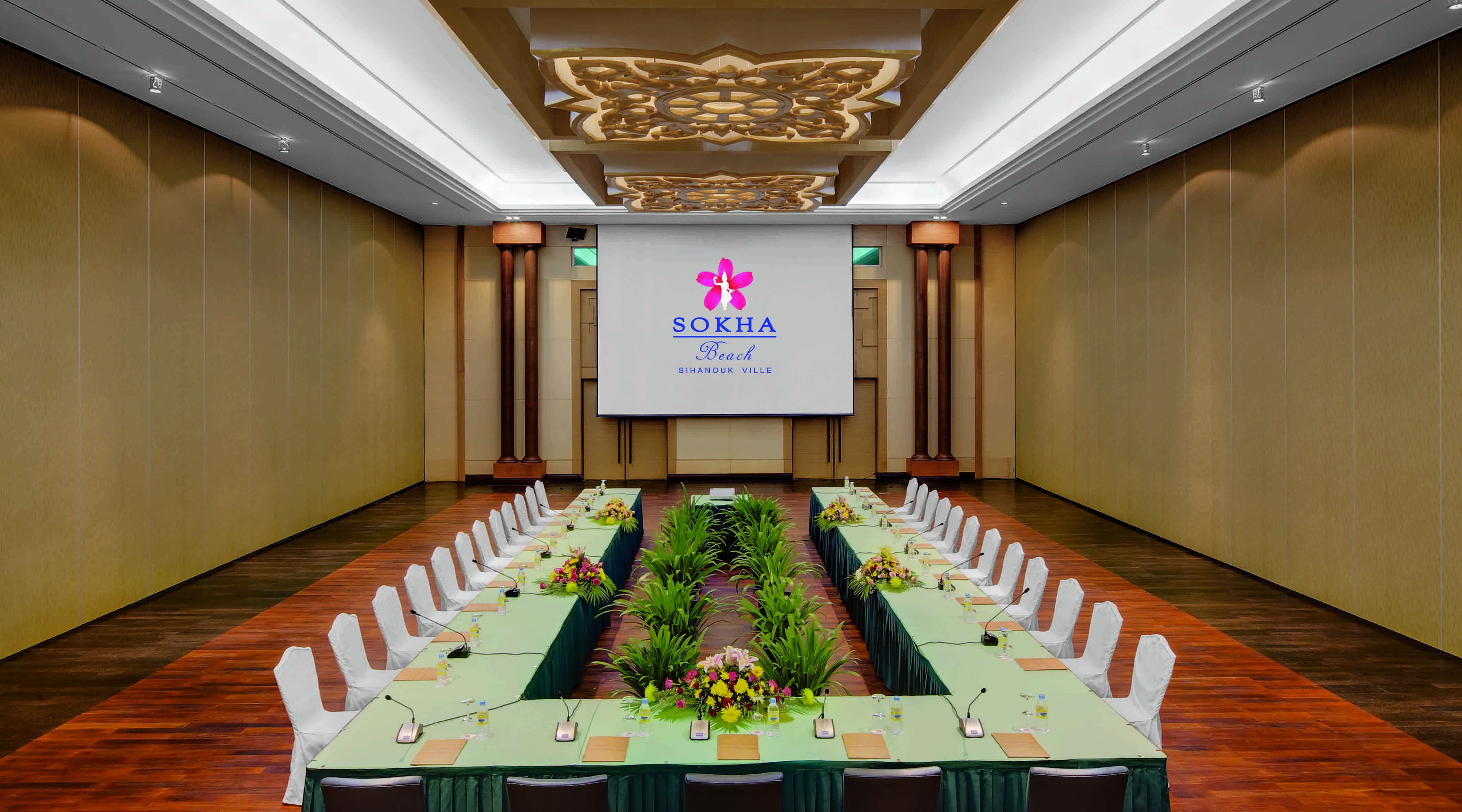 A conference room with a long table set for a meeting, a large screen displaying the logo 'SOKHA Beach Sihanouk Ville', and a symmetrical arrangement of chairs.