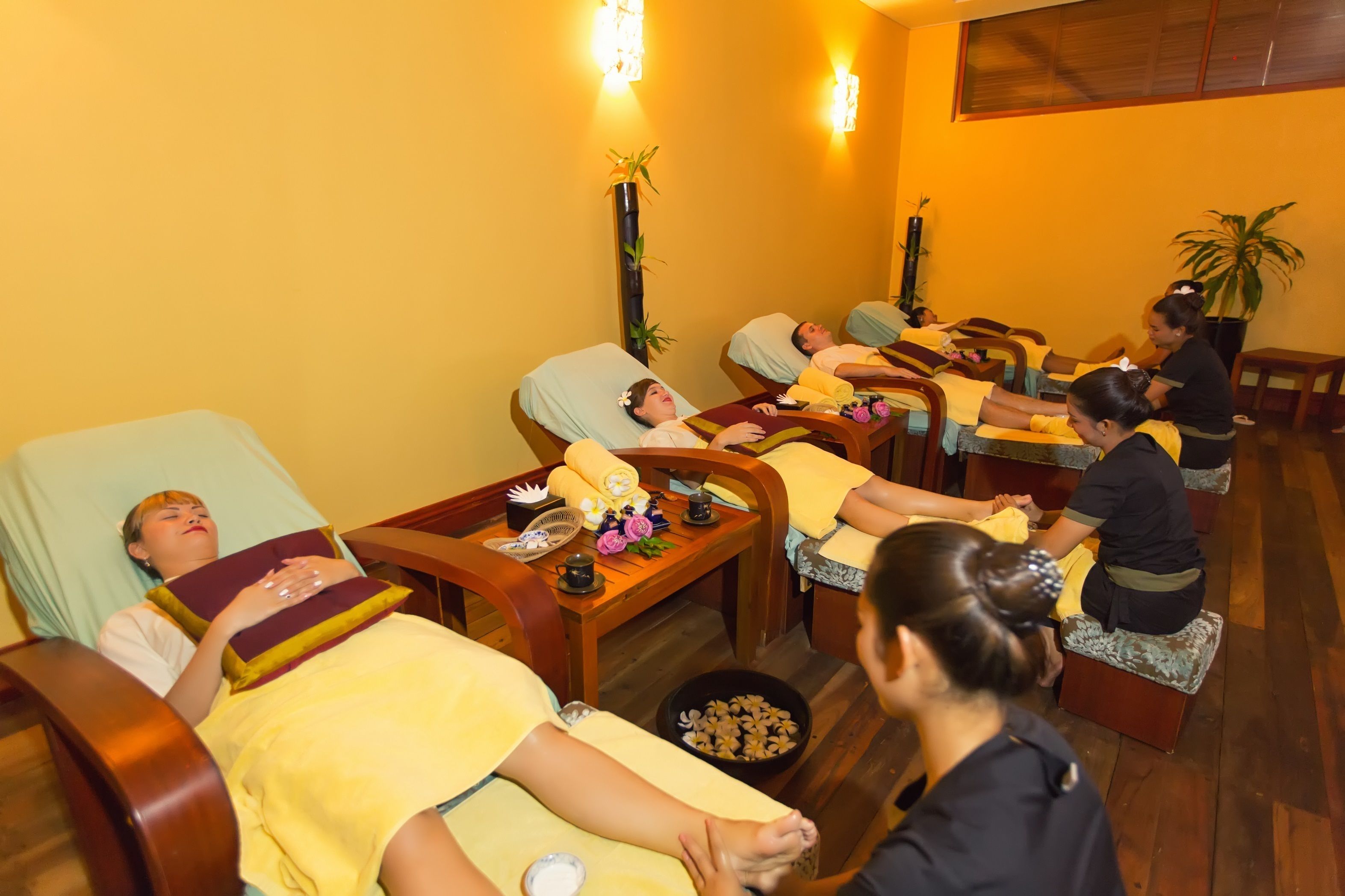 A spa room with multiple massage tables, each with a person lying down. There are attendants seated on stools, attending to the clients. The room has wooden flooring and walls, with decorative plants and a vase with flowers on a table.