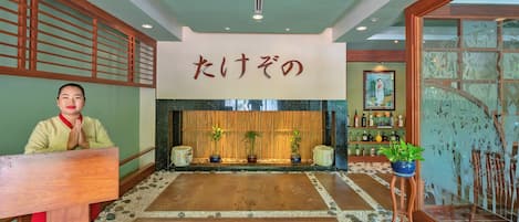 A traditional Japanese interior with a wooden reception desk, a sign with Japanese characters, a stone floor, and a bamboo screen.